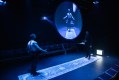 A stage awash in blue light. Two men face each other standing on a strip of carpet. A mirror disc hangs above and reflects one of them in this production of 'A Love Letter to the Nightingale.'