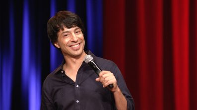 Arj Barker, a man with dark hair smiling. He is wearing a dark shirt and holding a microphone.