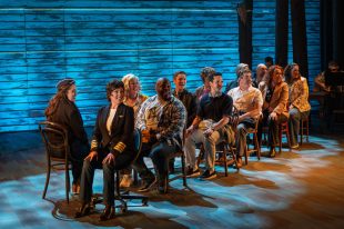 The cast of "Come From Away'. They are sitting in two rows of chairs, replicating the seats of an airplane.