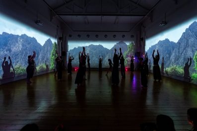 A group of performers are dancing in a gallery space. There are images of trees and mountains on screen panels around them.