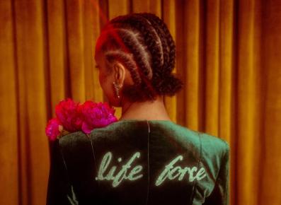 Singer Esperanza Spalding with her back turned to the camera. She has a jacket with the words 'life force' and is carrying some pink flowers. Her brown hair is styled in cornrows.