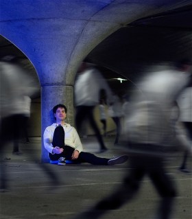 A young man is sitting and leaning against a pillar post. Variations of him are swirling around him in blurry forms.