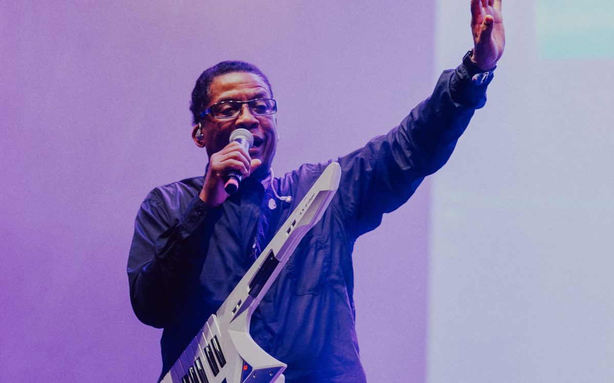 Herbie Hancock with a microphone to his mouth and a keyboard slung over his shoulder.