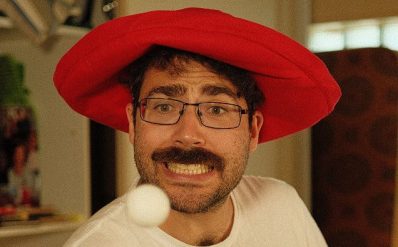 A brunette man, Jacob Sacher, with glasses and a moustache is wearing a floppy red hat. He's staring at a ping pong ball that's coming towards him. Jacob Sacher in Promising Young Mensch.