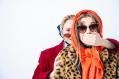 In a production of 'Years Years Bears', two women in colourful headscarves and sunglasses. One is standing behind the other and placing her hand over the other woman's mouth.