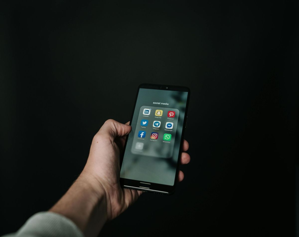 social media hacking: The hand of a white male person holding a smartphone displaying a range of social media icons on its screen against a black background.