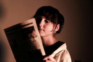 Woman with short dark hair reading newspaper in soft lighting. Arts news