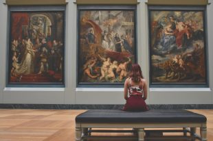 A young woman sits on a bench with her back to the camera, looking at three large classical paintings on a gallery wall.