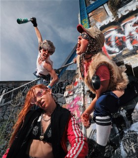 Three dancers, Oli Mathiesen, Lucy Lynch and Sharvon Mortimer, are dressed in rave gear and are posing and singing.