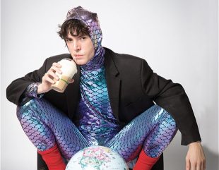 Elliot Wood dressed in a shimmery purple blue lizard costume, with a jacket over it. He is squatting in front of a globe and drinking from a straw.