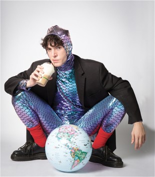 Elliot Wood dressed in a shimmery purple blue lizard costume, with a jacket over it. He is squatting in front of a globe and drinking from a straw.