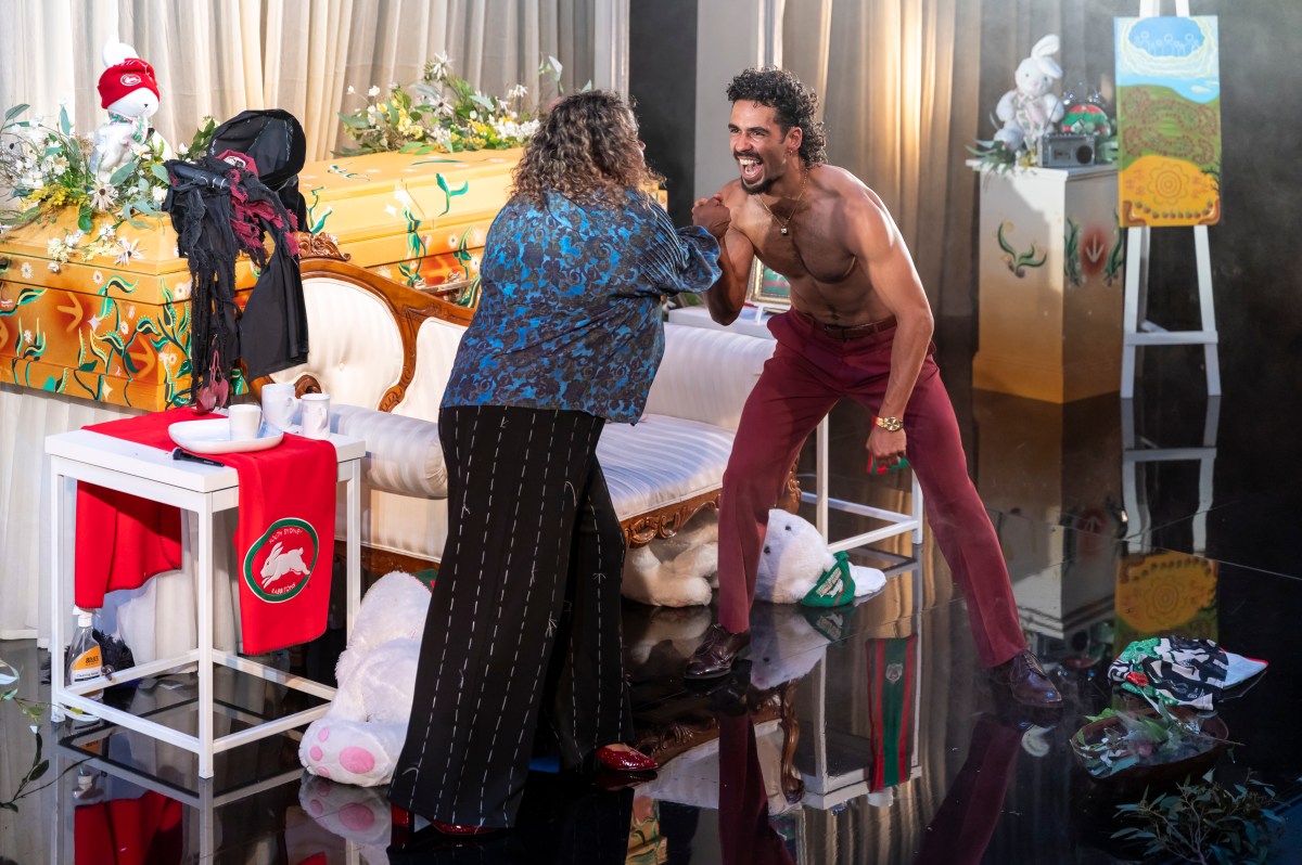 A First Nations woman in blue top and black pants shakes grabs the hand of a young bare-chested First Nations man, who has his mouth open wide smiling. They are surrounded by lots of colourful objects including a coffin. A Wake – A Woke Mob