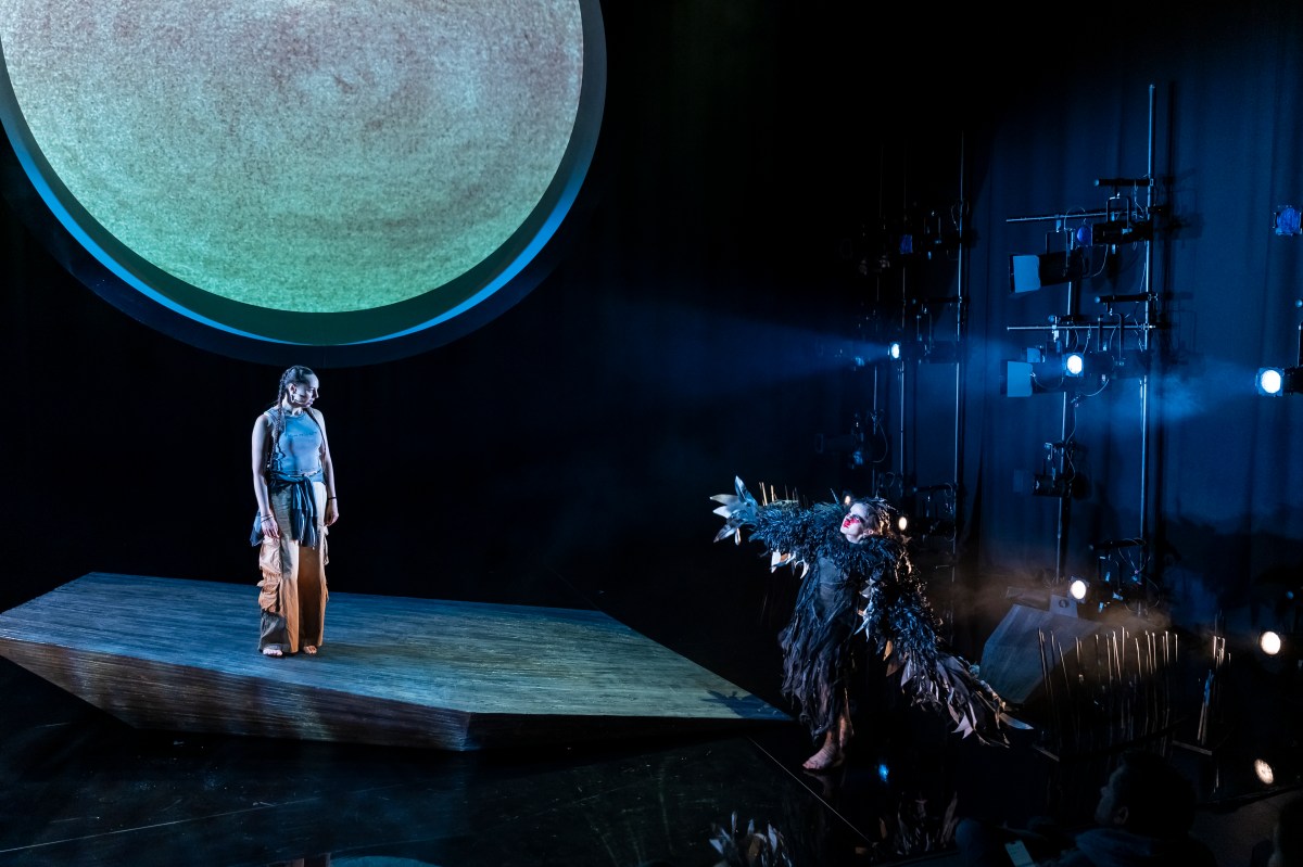 On a small geometric raked stage a young woman stands in traditional First Nations clothes under a large moon like set design, while an actor dressed like a crow dances. Gunawarra Re-creation.