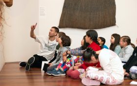 Participants in Culture Dose for Kids youth mental health program at the Art Gallery of New South Wales. A group of kids sitting on the floor of a gallery looking up as a guide points at an artwork.