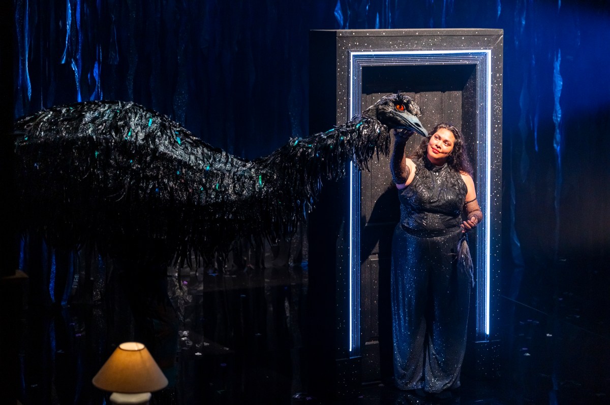 A young First Nations woman in a doorway strokes the head of a large human size emu puppet costume. Emu in the Sun.