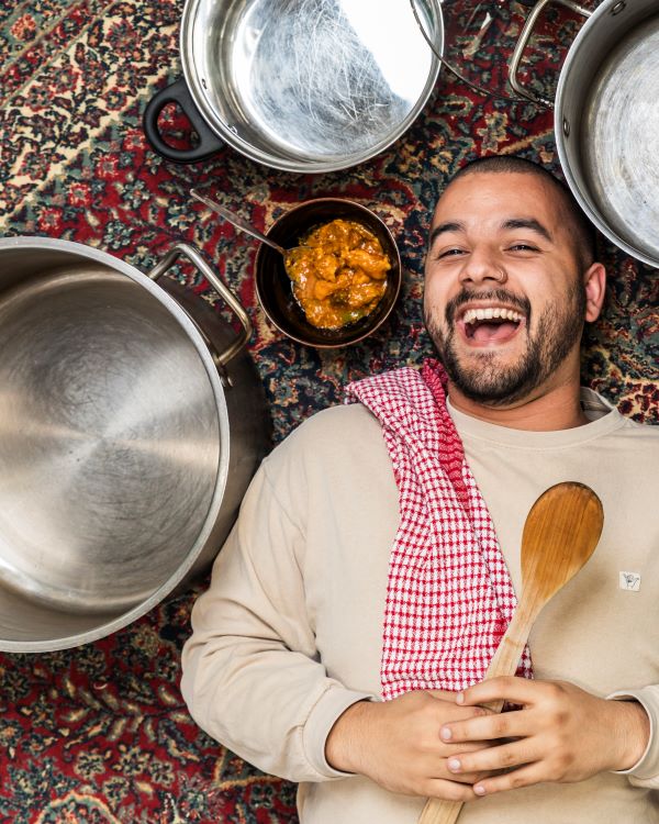 Josh Hinton's 'A Place in the Sultan’s Kitchen (or How To Make the Perfect One-Pot Chicken Curry)' receives its mainstage season at Merrigong Theatre Company in 2025. A bearded young man lies on the floor laughing. He is surrounded by cooking dishes and holds a wooden spoon.