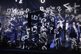 Four performers are barely visible as they overlaid by a scramble of letters.