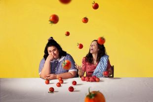 Brink Productions' 'Looking for Alibrandi' is sure to resonate with STCSA audiences. A young woman in a school uniform sits beside an older woman and a white cloth-covered table. Tomatoes sit on the table in front of them and rain down around them.