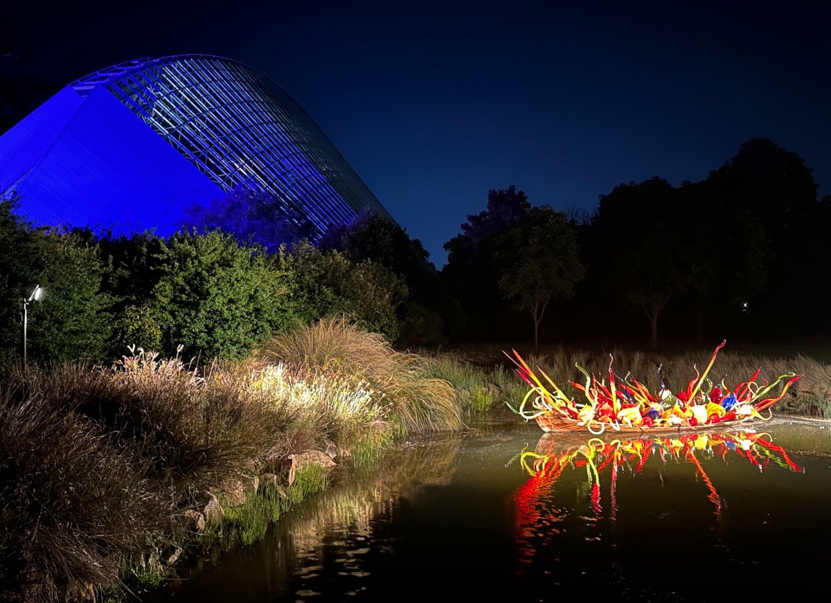 Night time view of an illuminated glass sculpture of a boat in garden setting. Dale Chihuly