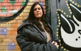 Claudia Gold aka Claw Money will debut in Australia with an exclusive exhibition at Warrnambool Art Gallery. A middle-aged woman with light brown skin wearing a black puffer jacket, standing in front of a brick wall with graffiti that is partially cropped.