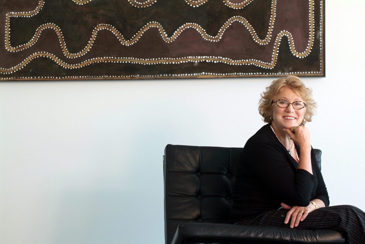 A middle aged Caucasian woman with short blond wavy hair and glasses sits on a chair on the right side of the picture with her chin resting on her hand. On the wall behind her is a piece of FN art. Private art collections.