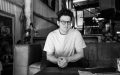 Queensland Theatre's new Artistic Director, Daniel Evans. A black and white photo of a young pale-skinned man with curly hair and round glasses sitting on a sofa in what appears to be a warehouse setting.