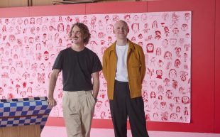 Two men, Dowel Jones, stand in pink decorated space. One wearing black top and light trousers, the other in white T shirt, brown jacket and dark trousers.