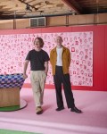 Two men, Dowel Jones, stand in pink decorated space. One wearing black top and light trousers, the other in white T shirt, brown jacket and dark trousers.