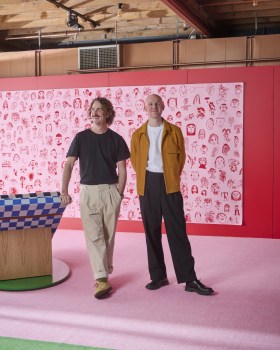 Two men, Dowel Jones, stand in pink decorated space. One wearing black top and light trousers, the other in white T shirt, brown jacket and dark trousers.