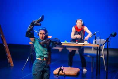 Two performers in 'Foleyvision'. Against a blue background one woman in the fore is wearing boots on her arms. The woman on the back is standing in front of a table.