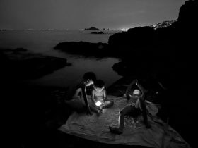 Black and white image of young boys at night lying on a rock near the sea with mobile phones with lit screens. Antonio Denti