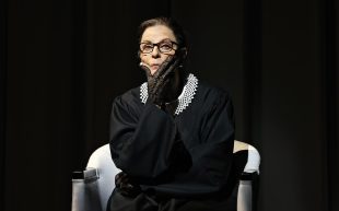 Heather Mitchell in Sydney Theatre Company’s ‘RBG: Of Many, One’ by Suzie Miller. A sharp-looking elderly woman dressed in black sitting on a white armchair with one of her hands framing her chin.