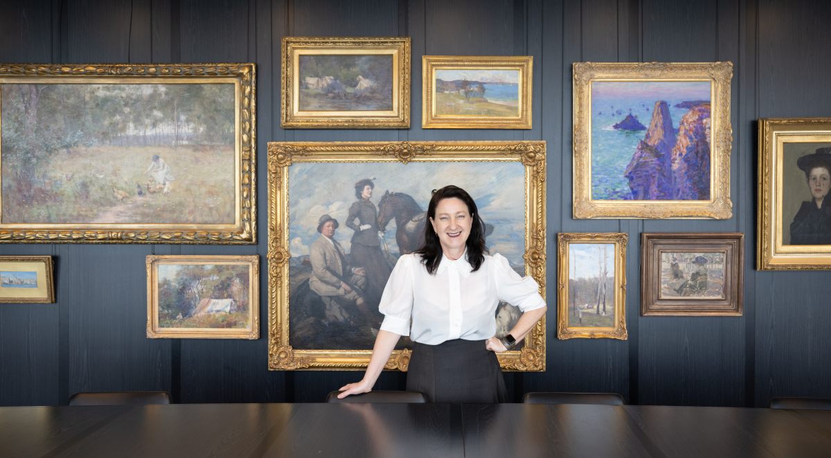 A Caucasian woman with dark shoulder length hair in a white shirt with dark skirt, stands behind a table with one hand on her hip and the other on the table. On the wall behind are a series of framed artworks. Private art collections