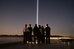 The light installation 'Karla Bidi' will run for the duration of Perth Festival 2025. Six people stand with their backs to the camera beneath a starry sky, on the banks of the Swan River. A bright beam of light rises from the riverbank into the sky before them.