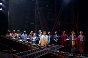 The cast of 'Jesus Christ Superstar'. A group are assembled around a central figure, a man. The is a large cross formation below them that also acts as a table.