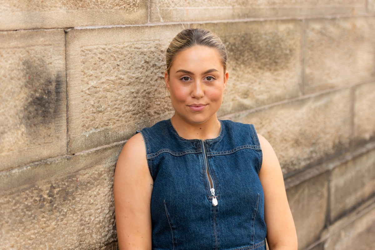 A young woman in a sleeveless blue denim dress with slicked back hair leans against a sandstone wall. National Arts School.