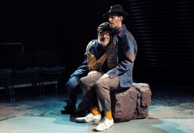 PJ Williams (left) as Estragon and Christopher Samuel Carroll (right) as Vadimir in The Street Theatre's 2024 production of 'Waiting for Godot'. A bearded man leans against another man wearing a hat, ad hugs him. They are sitting on a rock on stage; a park bench is visible behind them.