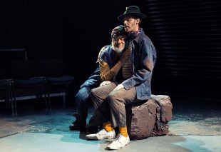 PJ Williams (left) as Estragon and Christopher Samuel Carroll (right) as Vadimir in The Street Theatre's 2024 production of 'Waiting for Godot'. A bearded man leans against another man wearing a hat, ad hugs him. They are sitting on a rock on stage; a park bench is visible behind them.