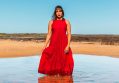 A publicity image for 'Yandha Djanbay (Go Slowly)' by Kirli Saunders, part of Merrigong Theatre Company's MERRIGONX program in 2025. A First Nations woman in a red dress stands ankle deep in a pool of water, an arid landscape and blue sky visible behind her.