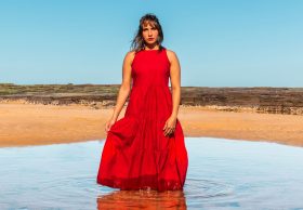 A publicity image for 'Yandha Djanbay (Go Slowly)' by Kirli Saunders, part of Merrigong Theatre Company's MERRIGONX program in 2025. A First Nations woman in a red dress stands ankle deep in a pool of water, an arid landscape and blue sky visible behind her.