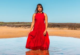 A publicity image for 'Yandha Djanbay (Go Slowly)' by Kirli Saunders, part of Merrigong Theatre Company's MERRIGONX program in 2025. A First Nations woman in a red dress stands ankle deep in a pool of water, an arid landscape and blue sky visible behind her.