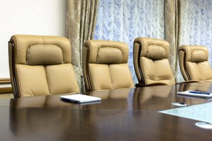 a row of empty padded beige chairs along a board room table. arts boards