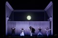 Five cast members of 'The Magic Flute'. They are dressed in shades of black, white and grey and are standing with their hands in the air. The grey set has an image of a moon above them.