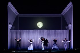 Five cast members of 'The Magic Flute'. They are dressed in shades of black, white and grey and are standing with their hands in the air. The grey set has an image of a moon above them.