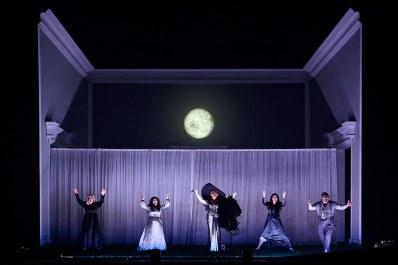 Five cast members of 'The Magic Flute'. They are dressed in shades of black, white and grey and are standing with their hands in the air. The grey set has an image of a moon above them.