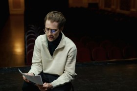 Man with cream sweater on dark theatre stage reading script for play.