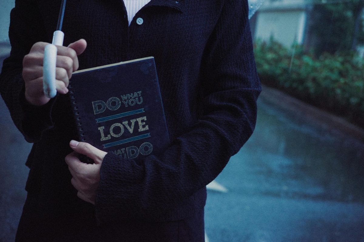 The torso and arms of someone in a dark jacket holding an umbrella and a book that says 'do what you love' on the cover. Self-publishing