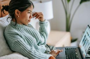 young Asian woman in light green sweater on couch looking at laptop computer. Arts news.