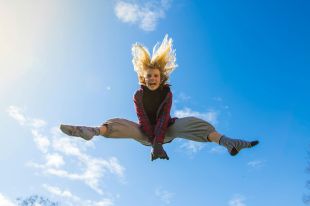 On the Move is ArtsHub's weekly round-up of arts sector appointments. The image shows a woman with long hair jumping in the aur. Her long hair flies up around her and her legs are outstretched. Blue sky and white clouds are visible behind her.