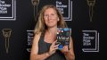 Author Samantha Harvey holding up her book 'Orbital'. She has long blonde hair and is wearing a black top. On the background is the Booker Prize 2024 signage.